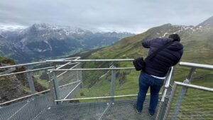 Visitante de costas na passarela de grade metálica da First Cliff Walk, suspensa sobre o vale de Grindelwald com montanhas ao fundo