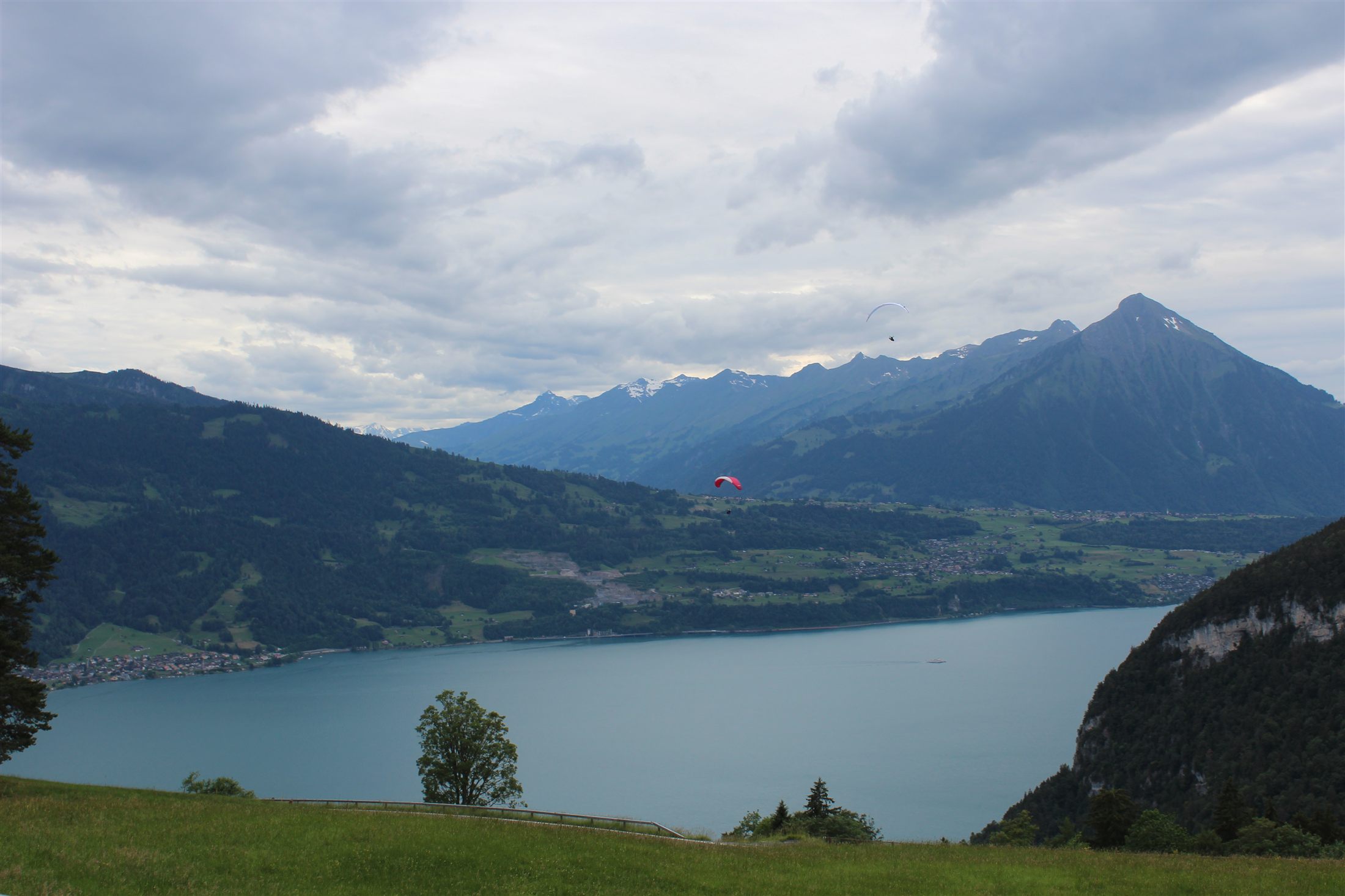 Parapente vermelho sobrevoando o vale aberto do Lago de Brienz, com caminho rural e montanha cônica à direita
