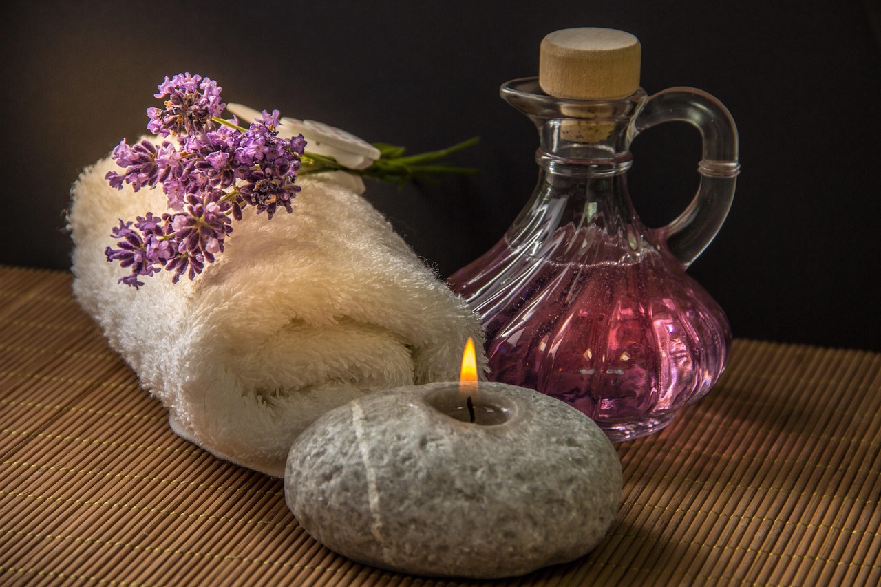 Toalha enrolada com flores de lavanda, um frasco de óleo e uma vela em pedra, o cenário de um ritual de spa