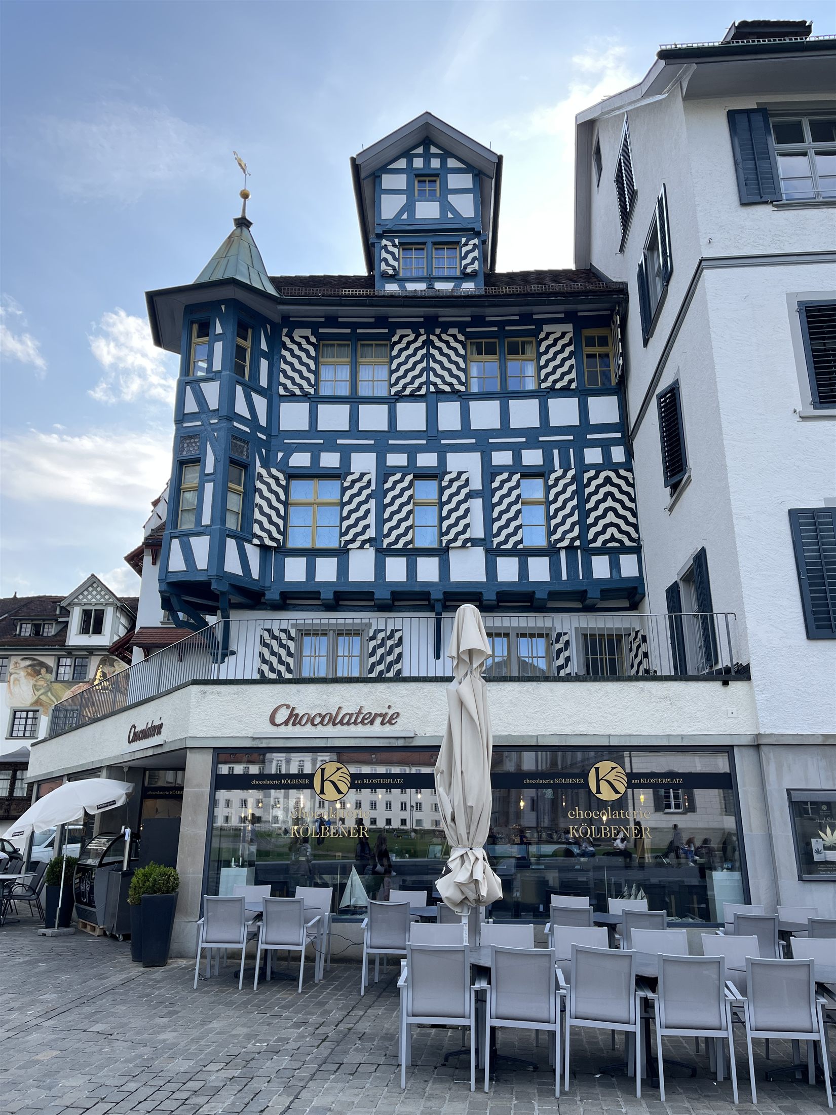 Fachada de uma chocolateria com edifício de enxaimel azul e branco ao fundo, na praça do mosteiro de São Galo