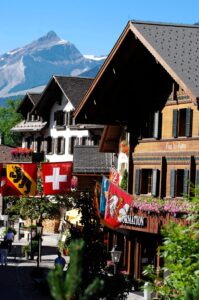 A Promenade de Gstaad no verão, com chalés de madeira entalhada, bandeiras suíças e do cantão de Berna e um cume alpino ao fundo