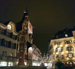 Fonte histórica iluminada no Marktplatz de Solothurn à noite, com o Zeitglockenturm e luzes de Advento ao fundo