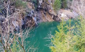 Água verde-turquesa entre paredões de rocha em camadas na Viamala, com mata de coníferas nas encostas, no inverno