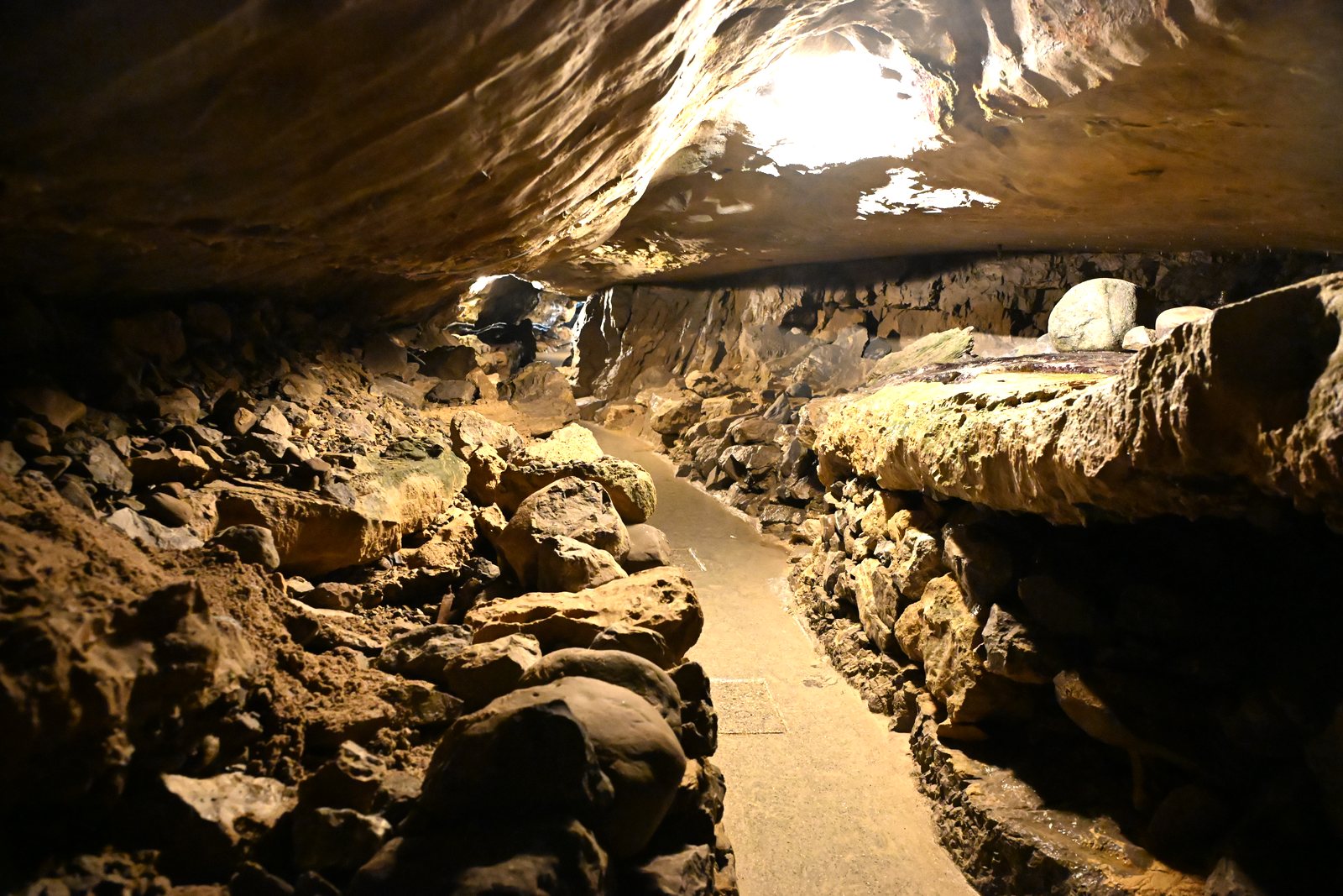 Caminho de concreto entre blocos de rocha desabados dentro das Grutas de São Beatus, com o teto iluminado por trás