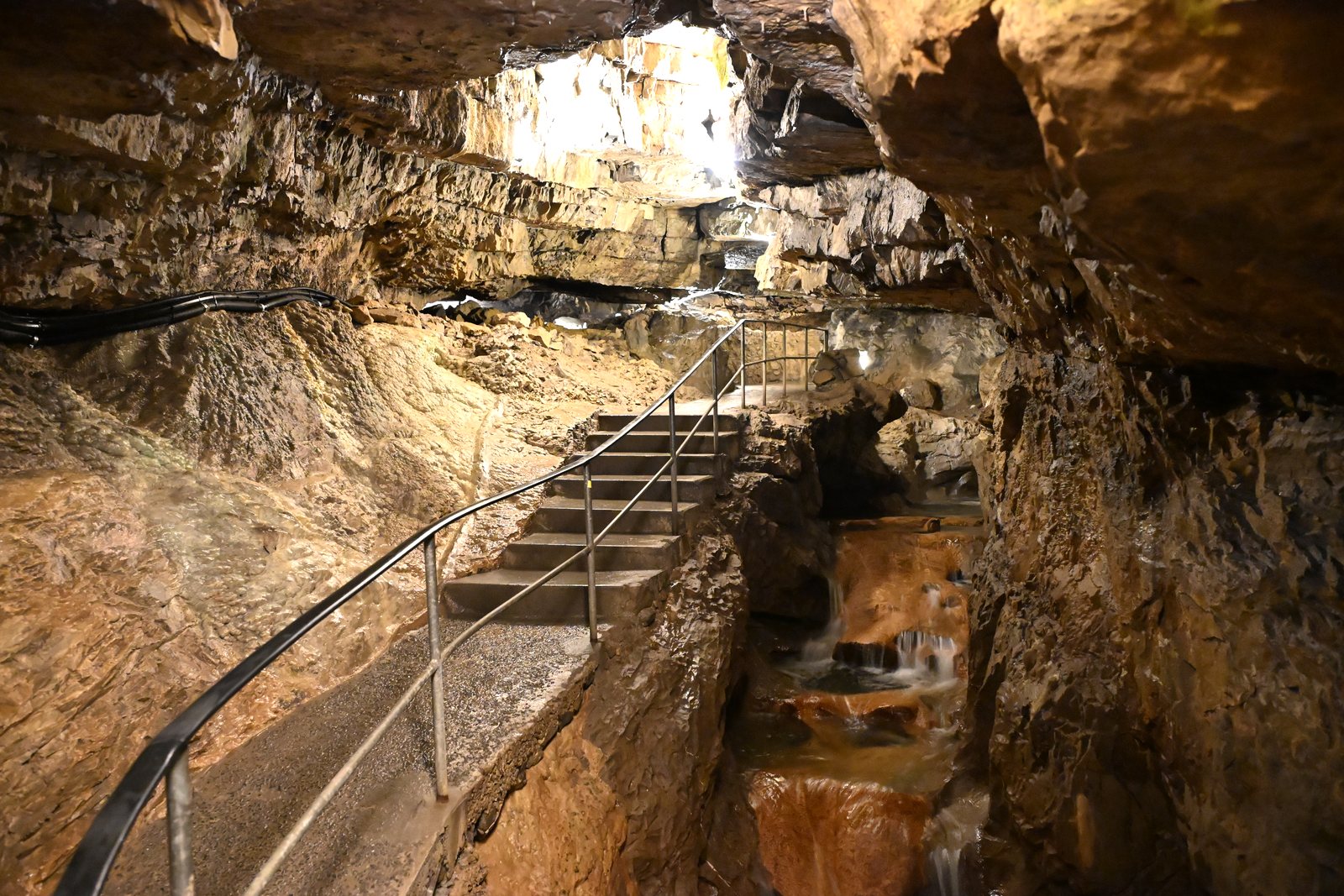 Escada e corrimão, ao lado da cascata, com uma abertura clara ao fundo