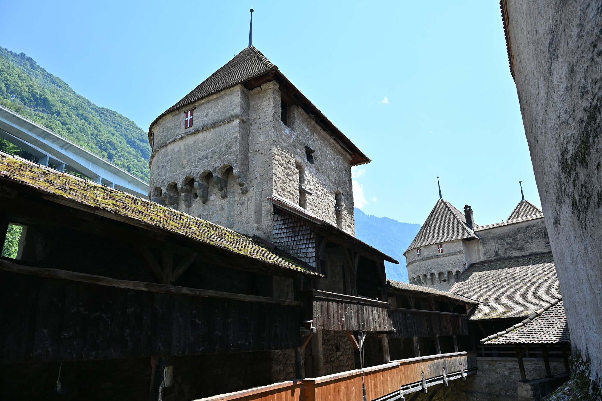 Adarves de madeira sobre a muralha do Castelo de Chillon do lado da montanha, com uma torre da cruz de Saboia e o viaduto da autoestrada ao fundo