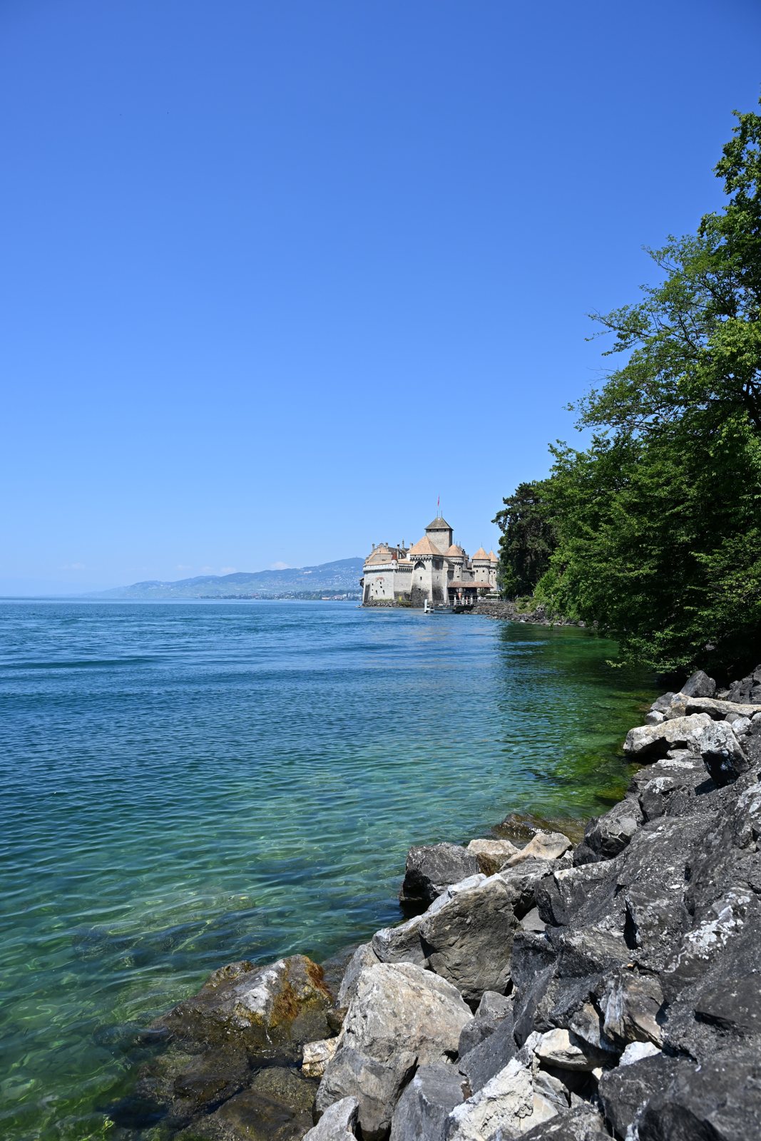 Castelo de Chillon visto da margem do Lago Léman, com a água verde e as pedras em primeiro plano