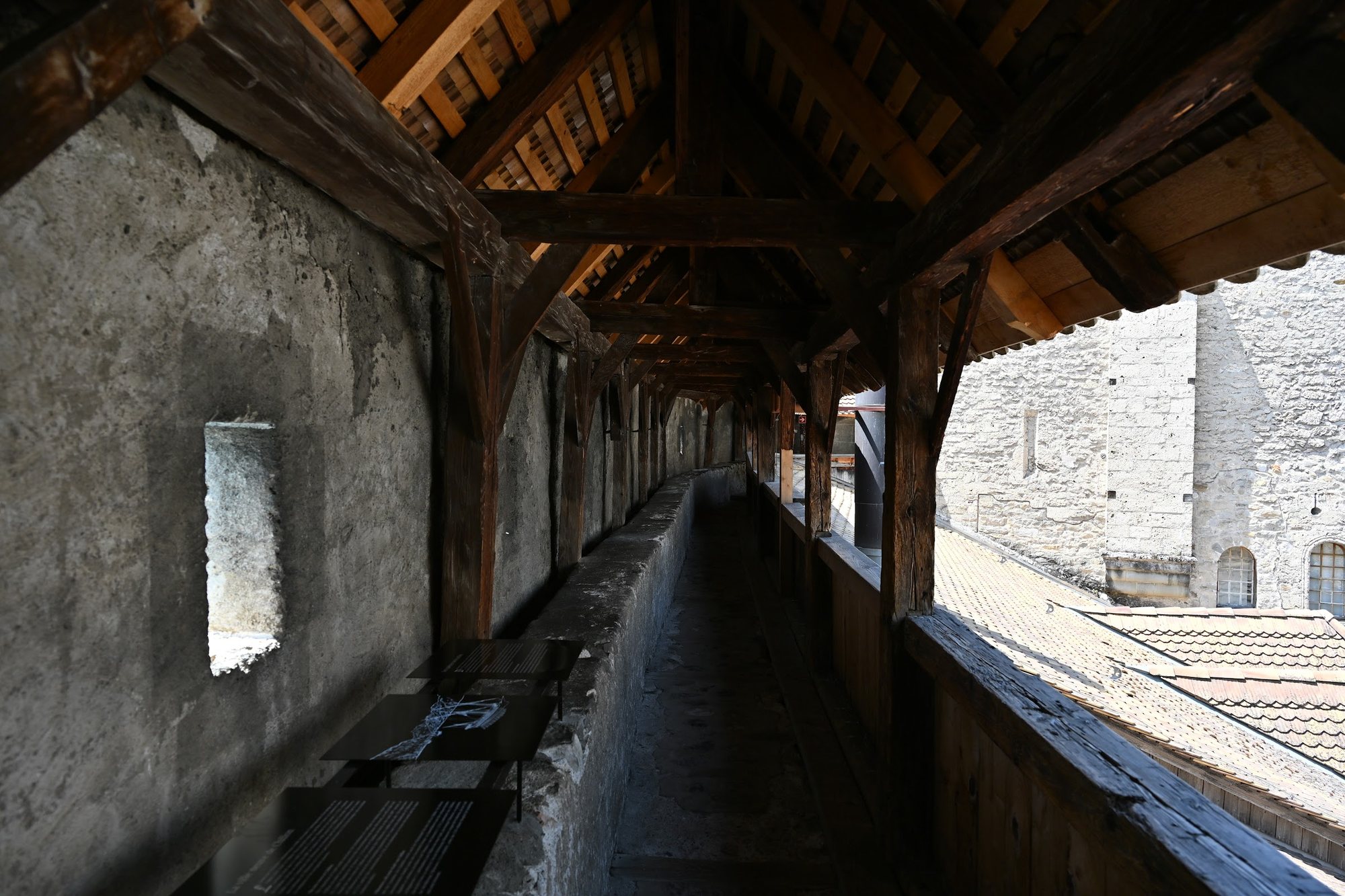 Interior do caminho de ronda coberto do Castelo de Chillon, com vigas de madeira e uma seteira aberta na parede de pedra