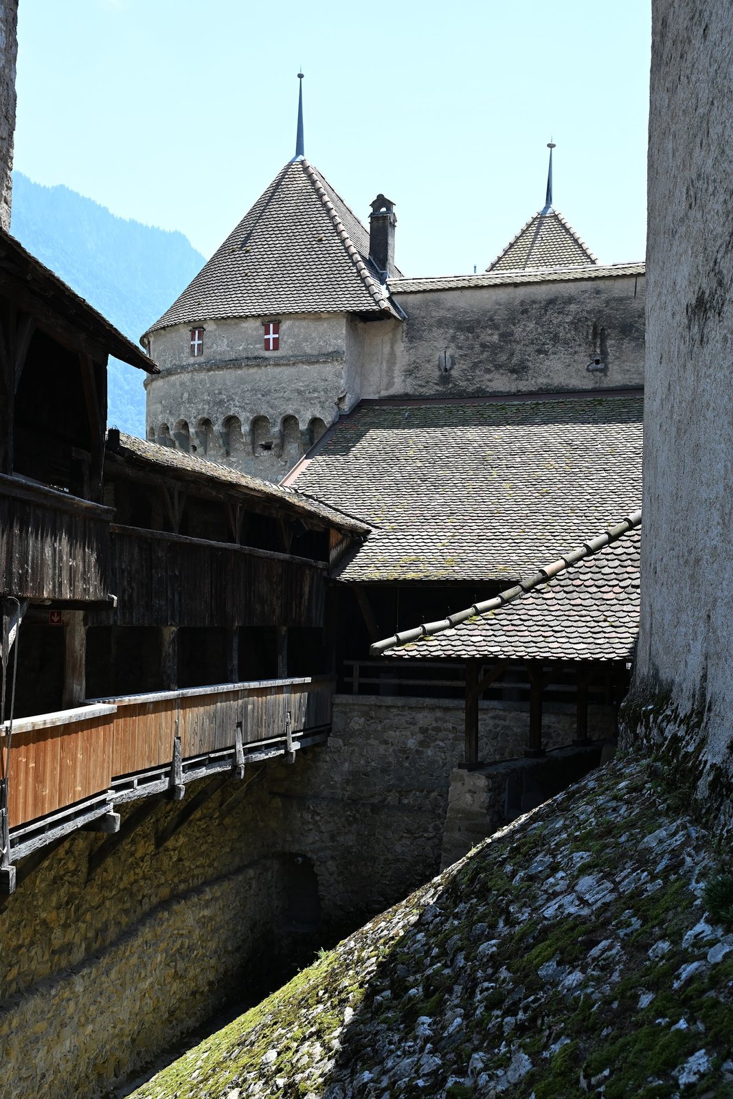 Rocha e muralha do Castelo de Chillon vistas do pátio da cortina, com a galeria de madeira do adarve à esquerda e uma torre redonda ao fundo