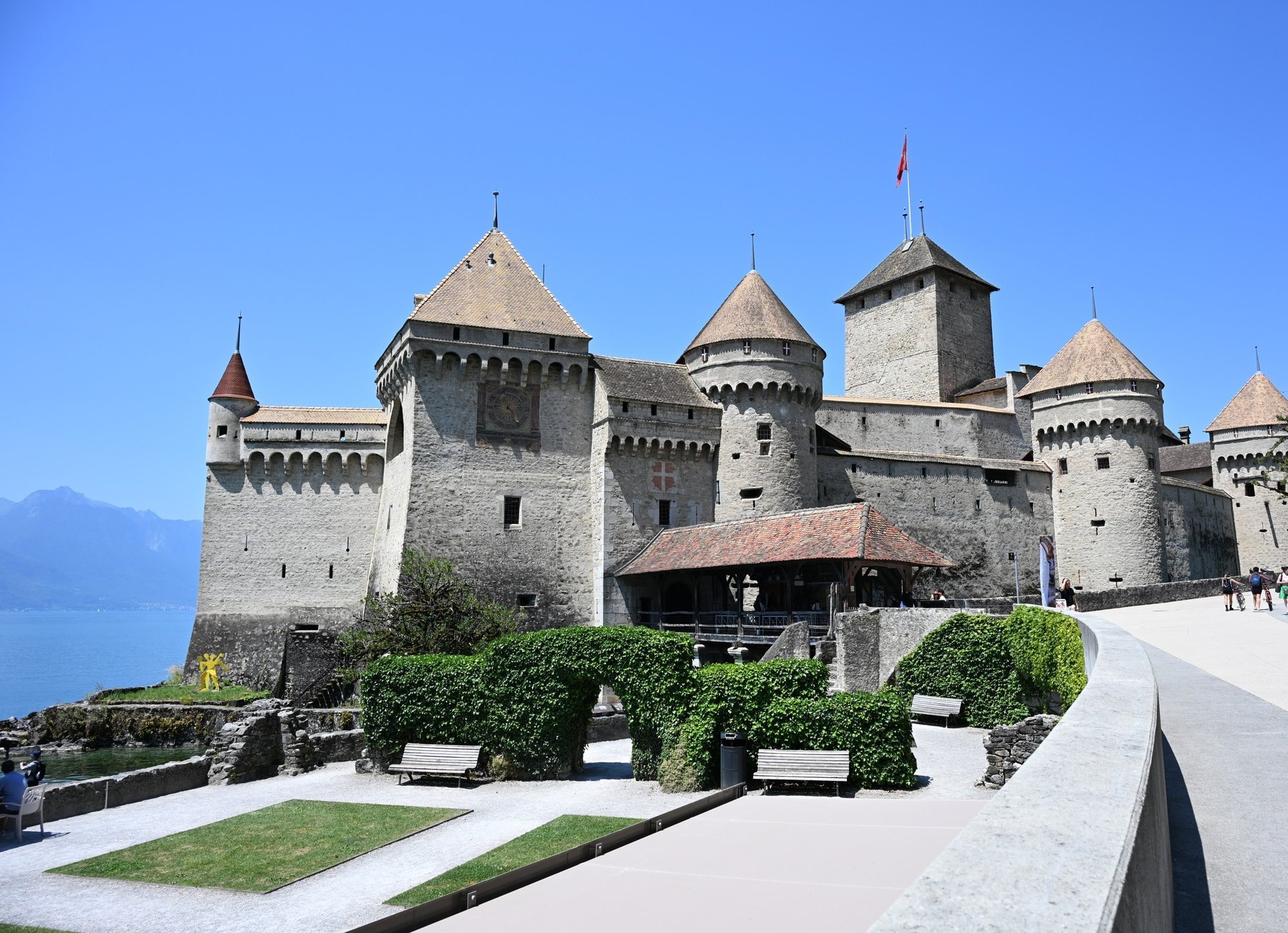 Frente do Castelo de Chillon com o jardim, a ponte coberta, a torre do relógio e a calçada larga da orla