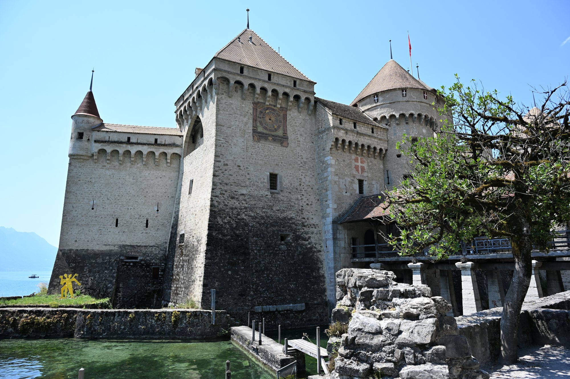 Torre do relógio e muralhas do Castelo de Chillon refletidas na água do fosso, com a bandeira suíça no torreão