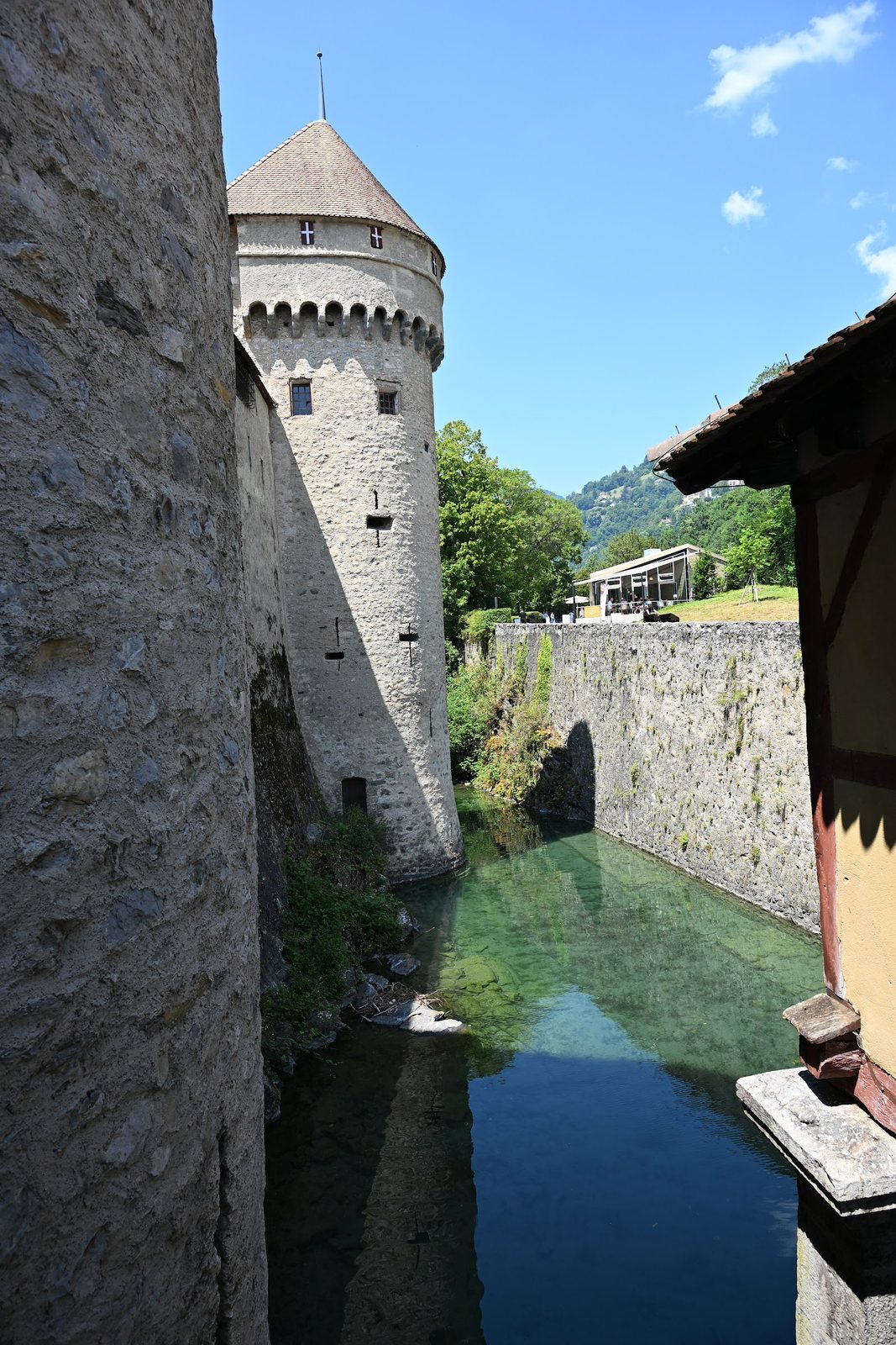 Fosso de água verde entre a rocha do Castelo de Chillon e a margem, com uma torre semicircular de matacães e seteiras em cruz
