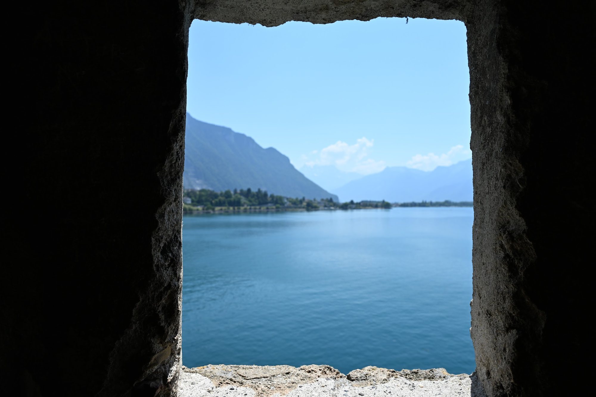 Janela de pedra do Castelo de Chillon emoldurando o Lago Léman azul, com a encosta de Villeneuve e as montanhas ao fundo