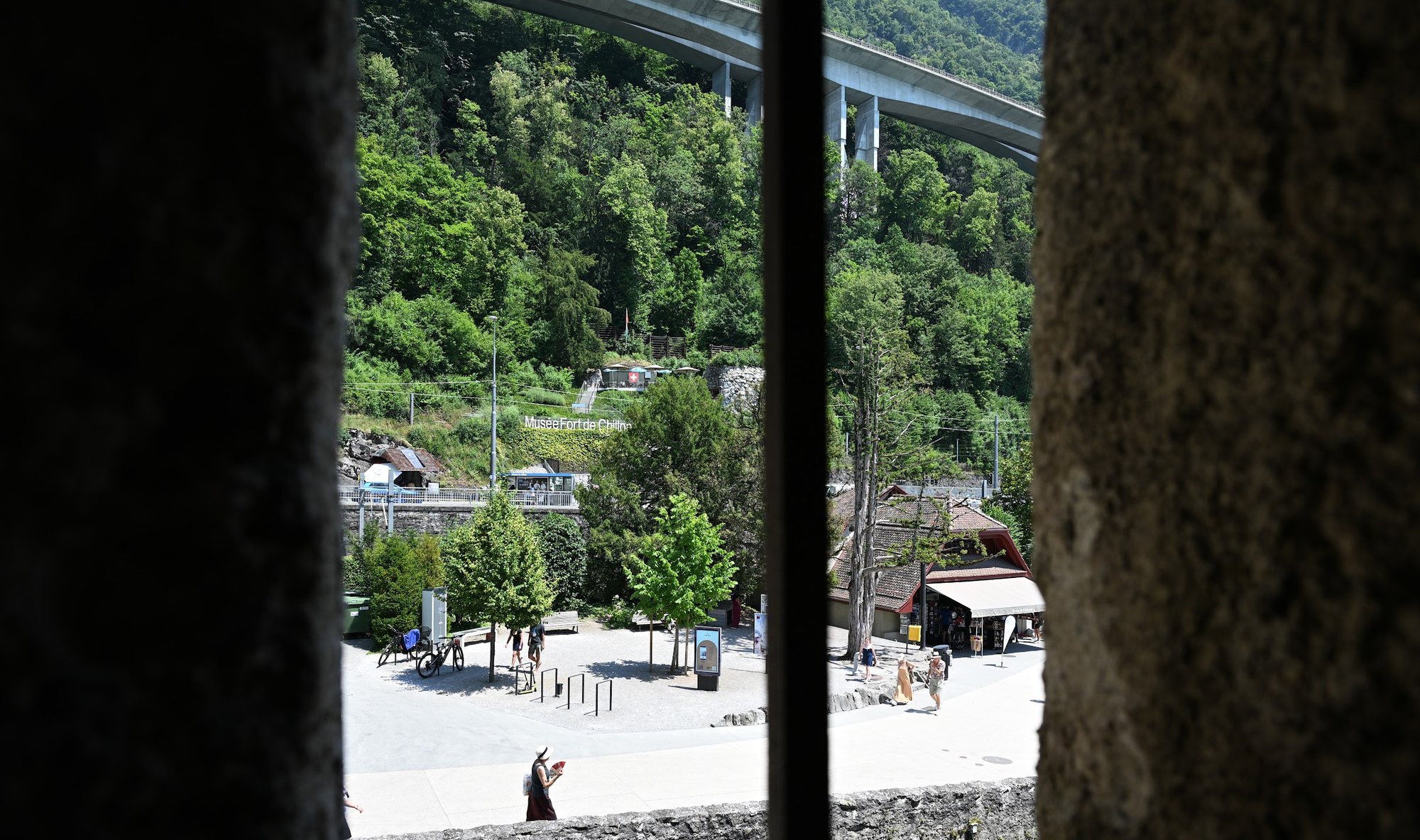 Vista por uma fresta do castelo para o largo da entrada, com o abrigo do ônibus, a placa do Musée Fort de Chillon e o viaduto da autoestrada ao fundo
