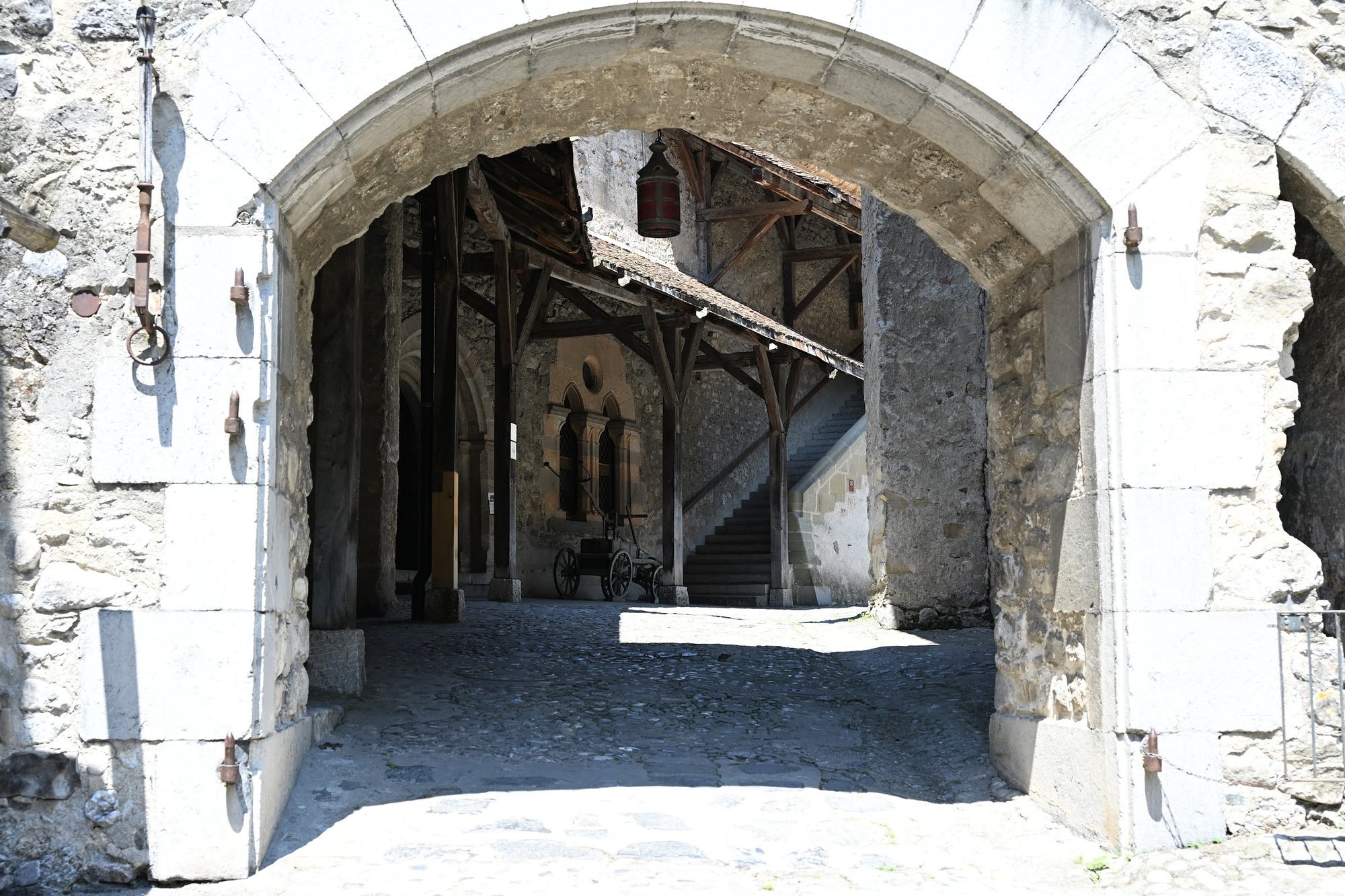 Portão em arco do Castelo de Chillon com argola e gonzos de ferro na pedra, e ao fundo o pátio com galeria de madeira e um carro de quatro rodas