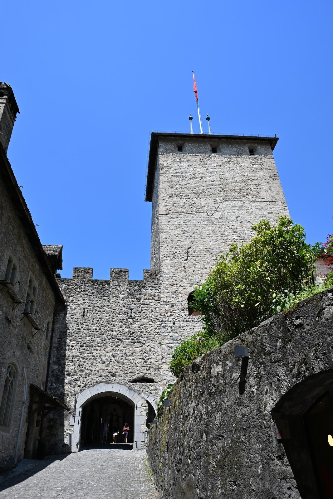 Rampa de pedra do Castelo de Chillon com muralha de ameias, duas seteiras e a torre de menagem quadrada erguendo-se ao fundo sob céu azul