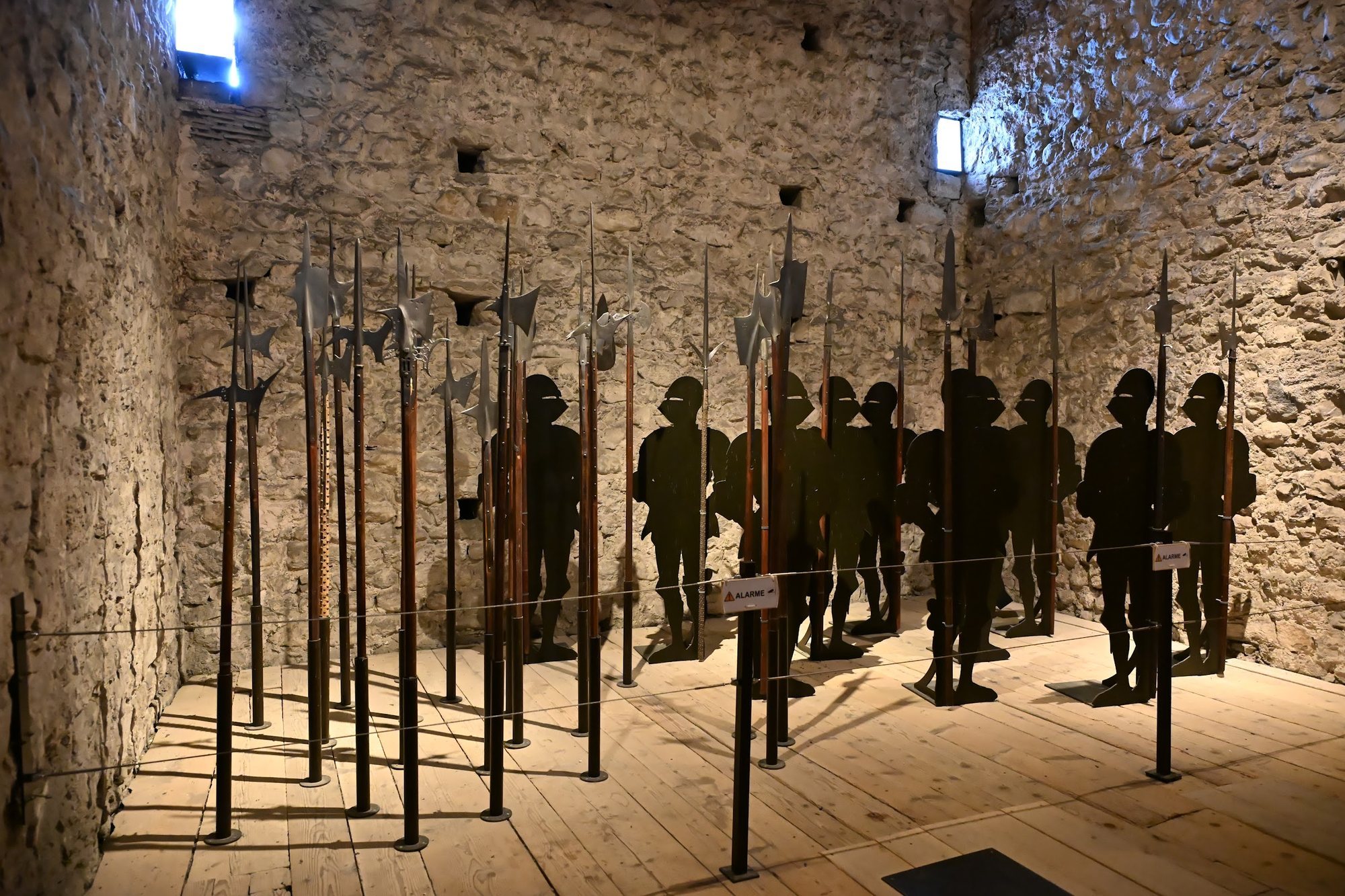 Sala de armas do Castelo de Chillon com alabardas e lanças em pé e silhuetas escuras de soldados ao fundo
