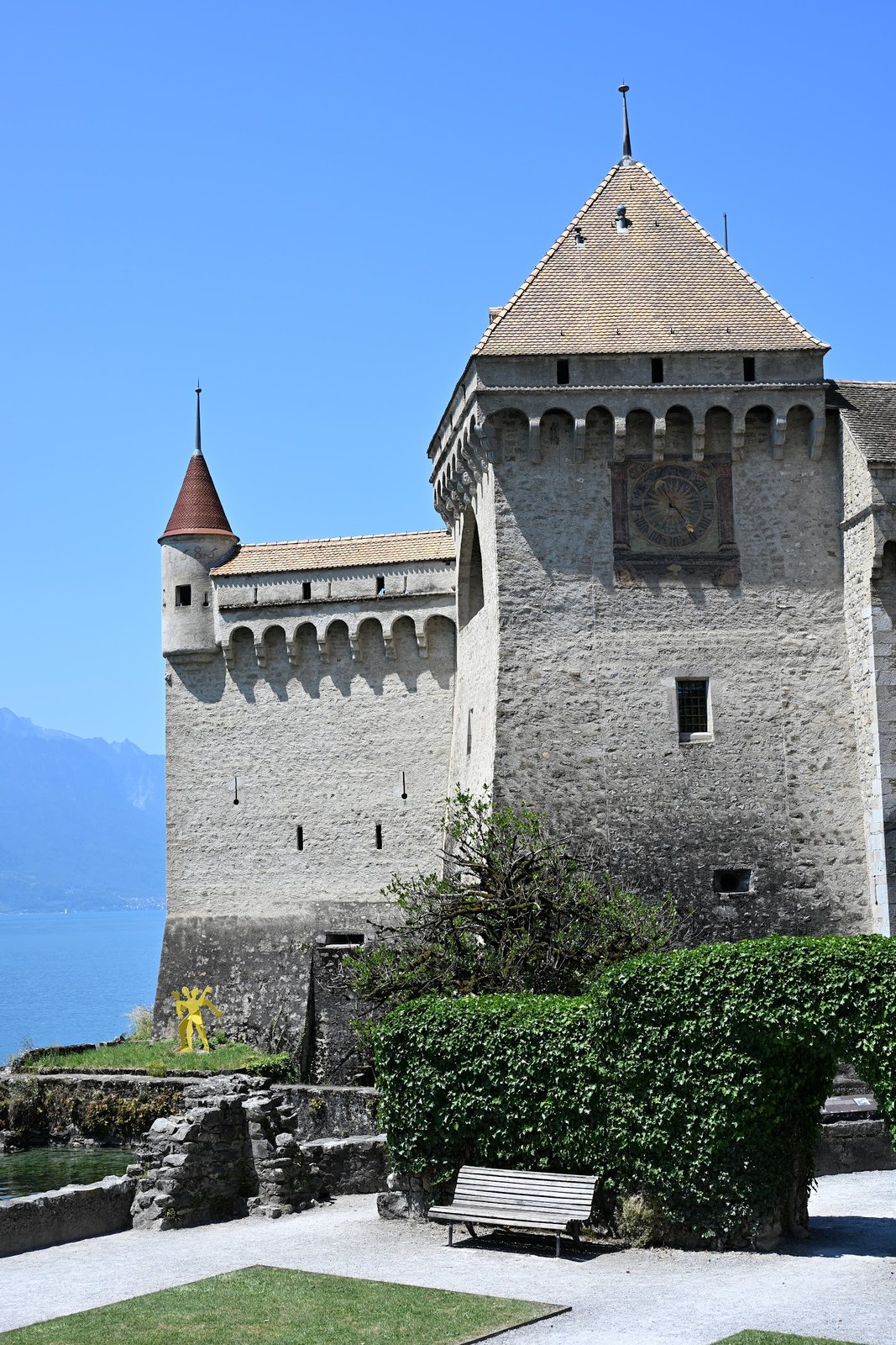 Torre de guarda do Castelo de Chillon com o relógio pintado, a fila de arcos dos matacães, a guarita de canto e a muralha com seteiras estreitas e aberturas em buraco de fechadura