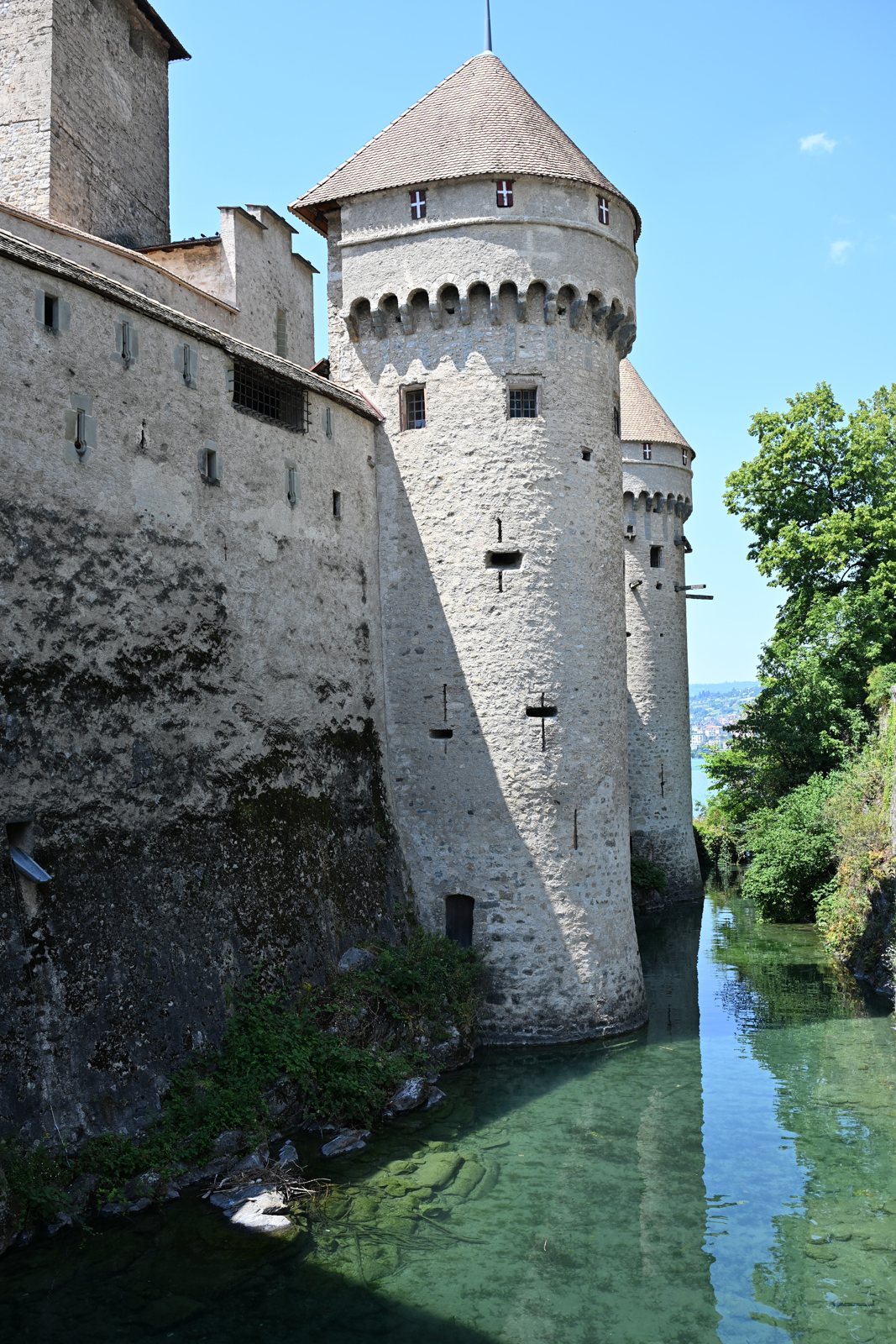 Torres redondas do Castelo de Chillon refletidas na água verde do fosso, com a cruz de Saboia pintada na pedra