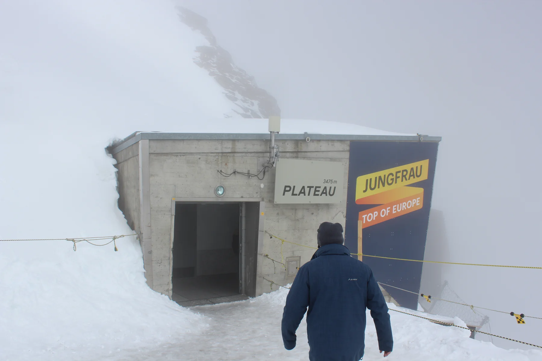 Entrada com placa Plateau entre neve e neblina, com um visitante de costas