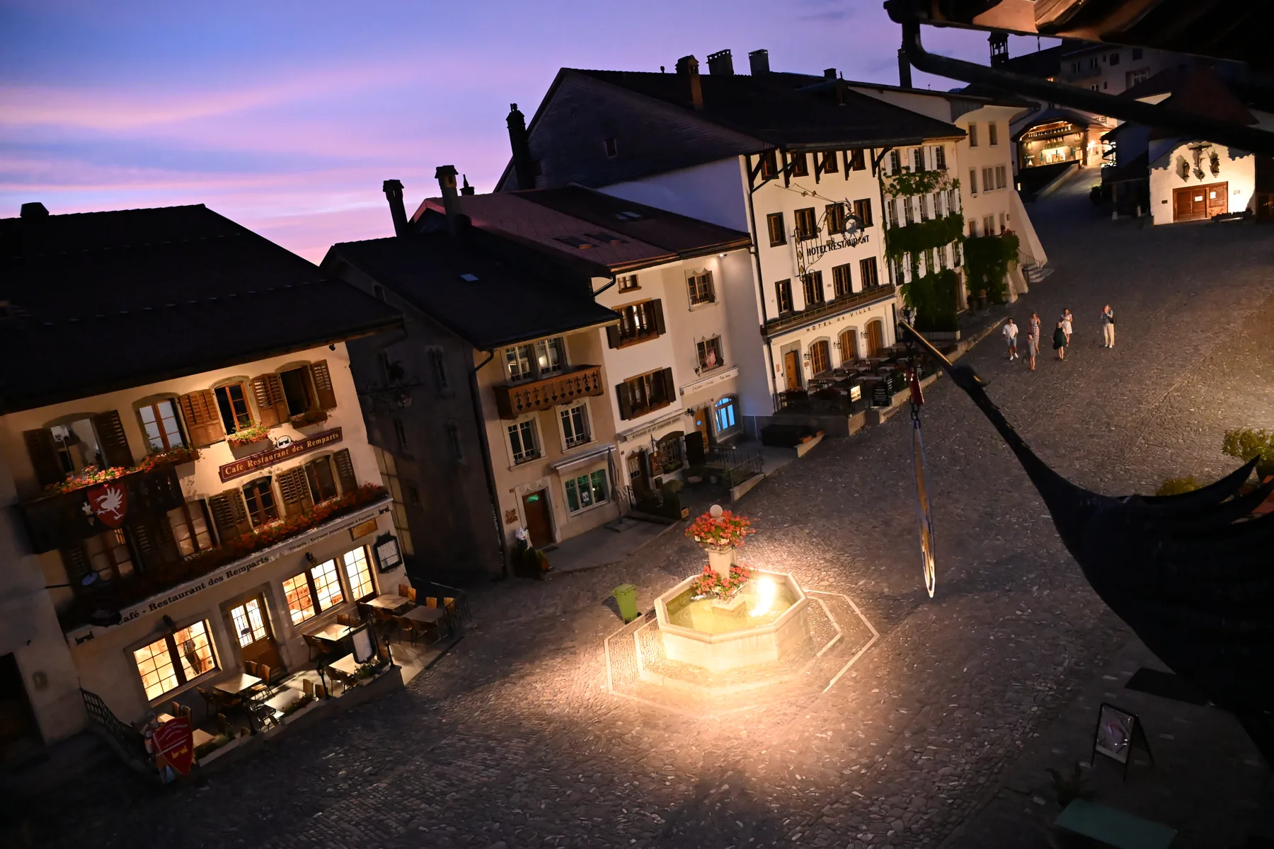 Rua de Gruyères ao anoitecer com fonte iluminada e fachadas